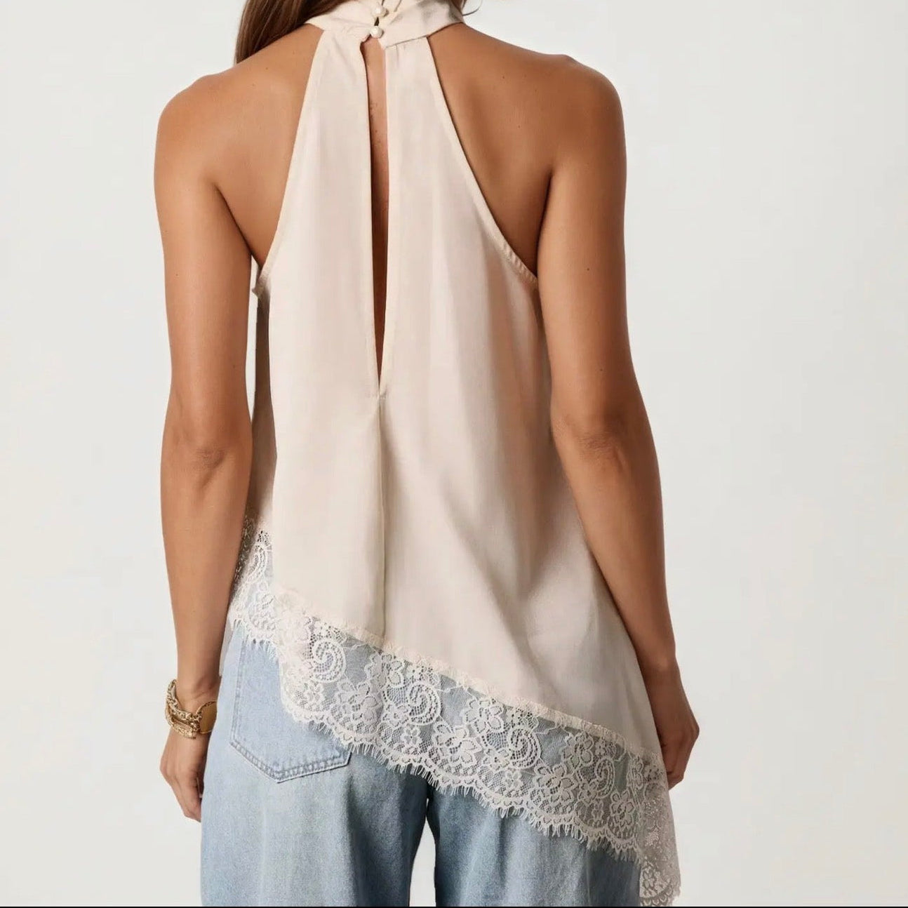 A woman shown from the back wearing a sleeveless cream halter top with a lace-trimmed hem and light blue jeans against a plain light background.