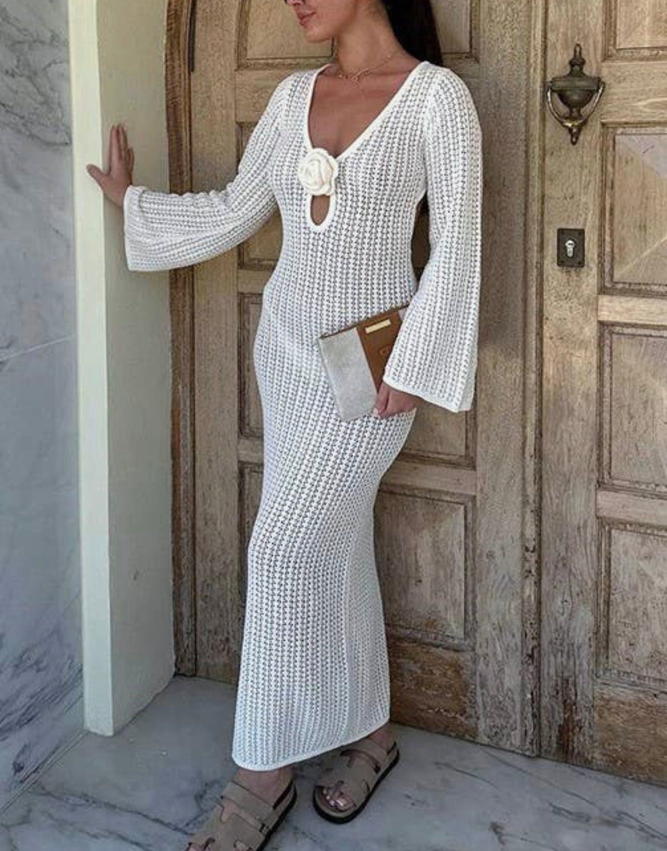 A woman models a white textured crochet maxi dress with long bell sleeves, a deep V-neckline, and a keyhole cutout at the chest. She is standing in front of a rustic wooden door and marble wall, holding a small brown clutch.